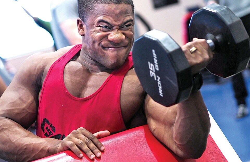 Man in a red tank top doing a preacher curl with his left hand, holding a 35 pound dumbell