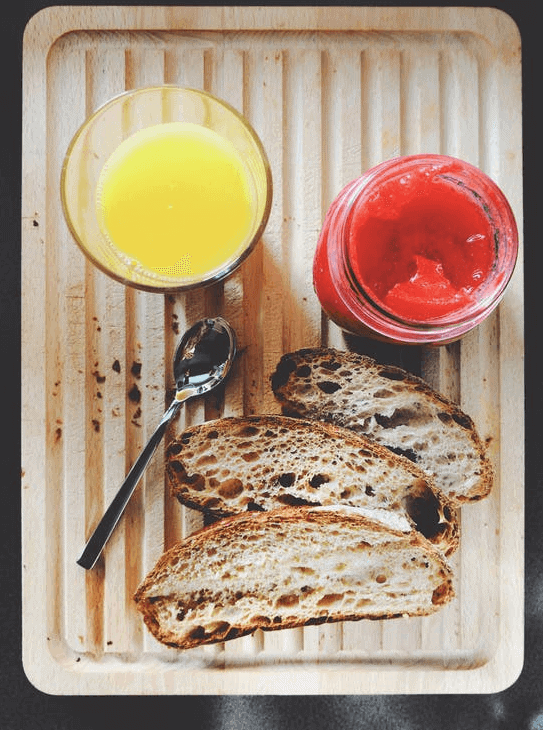 Glass of orange juice, strawberry jam, and toasted bread on top of a wooden board